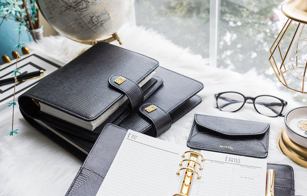 Black vegan leather planner laid open on a desk beside two other black planners.