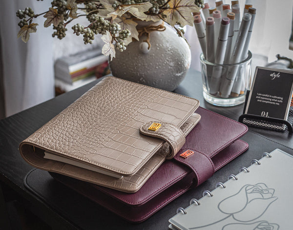 A beige croco planner on top of a mulberry saffiano planner atop a desk.