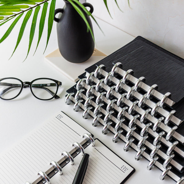 A stack of sleek black and silver discbound notebooks and a pair of glasses.